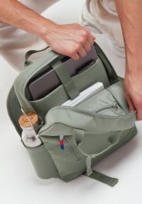 Zaino verde con finitura opaca, manico superiore, spallacci regolabili, tasca laterale che tiene una bottiglia d'acqua argentata e compartimento frontale con cerniera.