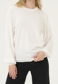 Vit stickad tröja med strukturerade ärmar, rundad halsringning och ribbad nederkant. Tillsammans med svarta byxor för kontrast.