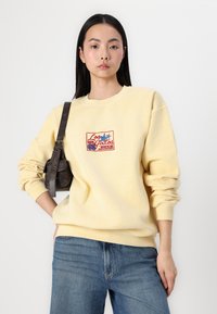 Gul sweatshirt med rundad halsringning och lös passform. Har en broderad röd och blå "Los Gatos"-design på bröstet. Bärs med jeans.