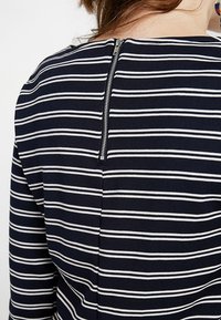 Mörkblå topp med vita horisontella ränder, med en dragkedja i ryggen och en slät, elastisk textur.