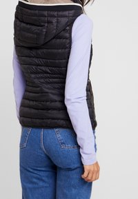 Czarna pikowana kamizelka z kapturem, z ozdobnym przeszyciem i pikowaną fakturą. Noszona na jasnofioletowej bluzce z długim rękawem i dżinsach.