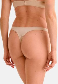 String beige en matière lisse, offrant une couverture minimale et des bretelles latérales fines, vu de l'arrière.