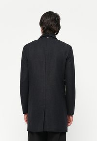 Un homme aux cheveux sombres et ondulés porte un long manteau en charbon sombre, se tenant de dos devant un fond blanc uni.