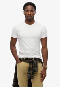 Biała koszulka z okrągłym dekoltem wykonana z bawełny, noszona z khaki spodniami oraz zieloną koszulą w kratę związaną w talii. Posiada małe logo na klatce piersiowej.