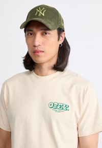 Camiseta beige con logo verde "OBEY" en el lado izquierdo del pecho; gorra de pana oliva con logo bordado "NY". El modelo tiene el pelo largo y lleva un pendiente.