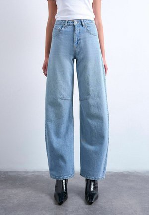 Personne portant un jean bleu clair à jambes larges et des bottines noires à bouts pointus, debout sur un sol gris face à un mur blanc uni.