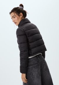 Žena s viazanými vlasmi má na sebe skrátenú čiernu puffovú bundu a tmavé džínsy s vysokým pásom a vreckami v tvare hviezdy na zadnej strane, pózuje bokom.