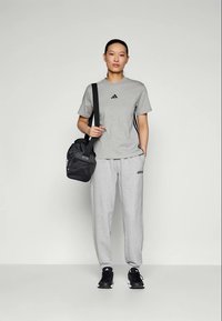 Γκρι μπλούζα Adidas με μαύρο λογότυπο, σε συνδυασμό με ανοιχτό γκρι παντελόνι φόρμας με λογότυπο, μαύρα αθλητικά παπούτσια και μια μαύρη τσάντα ώμου.