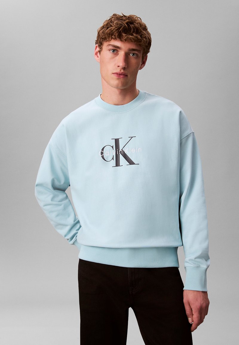 Jeune homme aux cheveux bouclés portant un sweat-shirt Calvin Klein bleu clair et un pantalon noir, debout devant un fond gris uni.