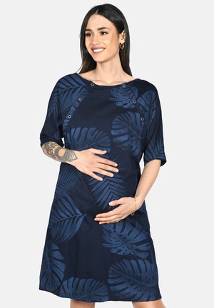 Smiling pregnant woman with tattoo on forearm wearing navy blue dress with large leaf patterns, hands cradling belly.