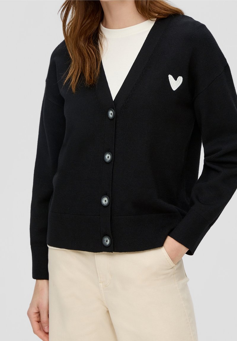 Femme portant un cardigan noir boutonné avec un détail cœur blanc sur un top blanc et un pantalon beige, une main dans la poche.
