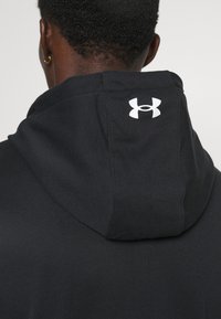 Vista posteriore di una persona che indossa un felpa nera con il logo bianco di Under Armour sul cappuccio, con capelli corti e ricci e orecchini a cerchio dorati.