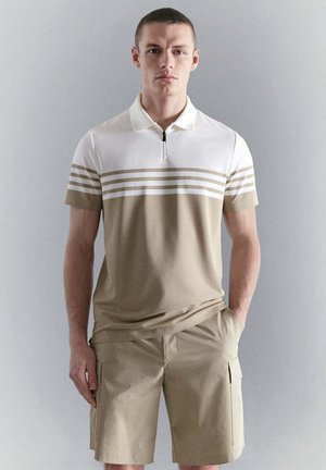 Jeune homme debout contre un mur gris clair, portant un polo beige et blanc à manches courtes et un short cargo beige, les mains dans les poches.