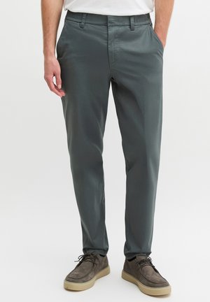 Mann trägt graue, schlanke Hose mit seitlichen und hinteren Taschen, kombiniert mit grauen Wildleder-Schnürschuhen mit hellbeigen Sohlen.