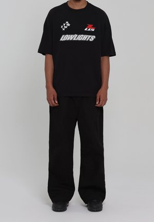Person trägt ein schwarzes übergroßes T-Shirt mit weißen und roten Logos über dem Wort "LOWLIGHTS", kombiniert mit lockeren schwarzen Hosen und schwarzen Schuhen.