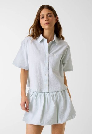 Jonge vrouw in lichtblauw, gestructureerd kort mouwen overhemd met knopen en bijpassende geplooide minirok tegen een effen witte achtergrond.