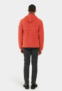 Orange fleecejacka med huva, textil med struktur, kontrasterande sömmar och detalj i ryggen. Styler med svarta jeans och stövlar.