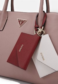 Różowa teksturowana torebka z logo Guess. Zawiera przymocowany złotym pierścieniem czerwono-biały uchwyt na karty. Gładkie wykończenie ze skóry.