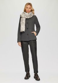 Manteau en mélange de laine gris au design ajusté, avec un col haut et des boutons. Associé à un pantalon en cuir noir et une écharpe légère à motifs.