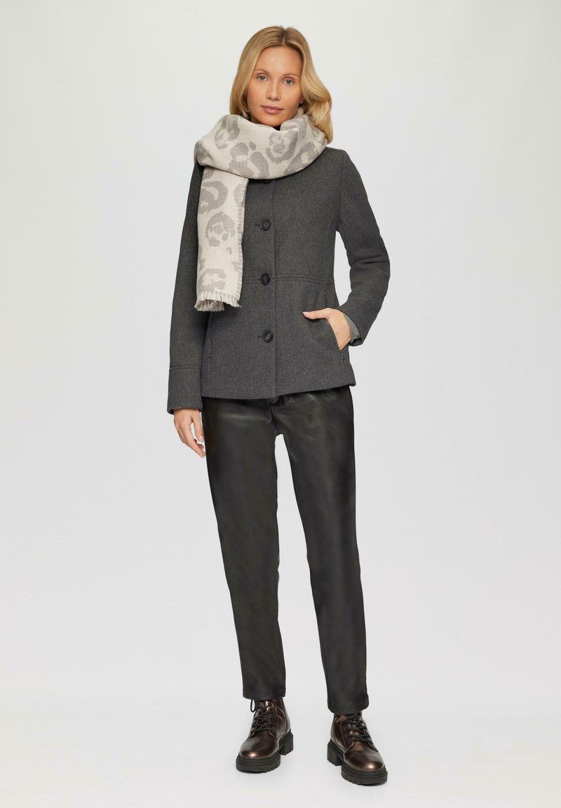 Manteau en mélange de laine gris au design ajusté, avec un col haut et des boutons. Associé à un pantalon en cuir noir et une écharpe légère à motifs.