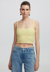 Top amarillo corto con un acabado texturizado, que presenta tirantes delgados y un escote cuadrado, combinado con jeans de mezclilla azul claro.