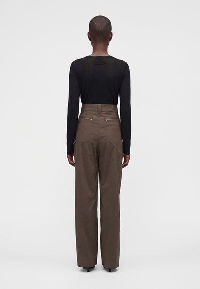 Personne aux cheveux courts portant un pantalon marron taille haute et un haut noir moulant à manches longues, debout de dos sur un fond uni.