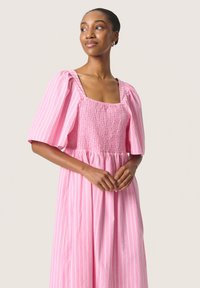 Rosa randig klänning med fyrkantig halsringning, puffärmar och smockad midja. Har en flödande kjol och lätt material.