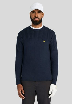 Mand iført en navyblå sweater med et gult logo, sorte bukser, hvid kasket og hvid golfhandske, stående mod en ensfarvet grå baggrund.