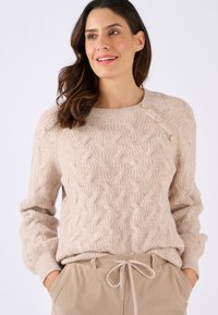 Pull en tricot beige avec un motif en câble, col rond et trois boutons décoratifs à l'épaule ; associé à un pantalon en velours côtelé beige clair.
