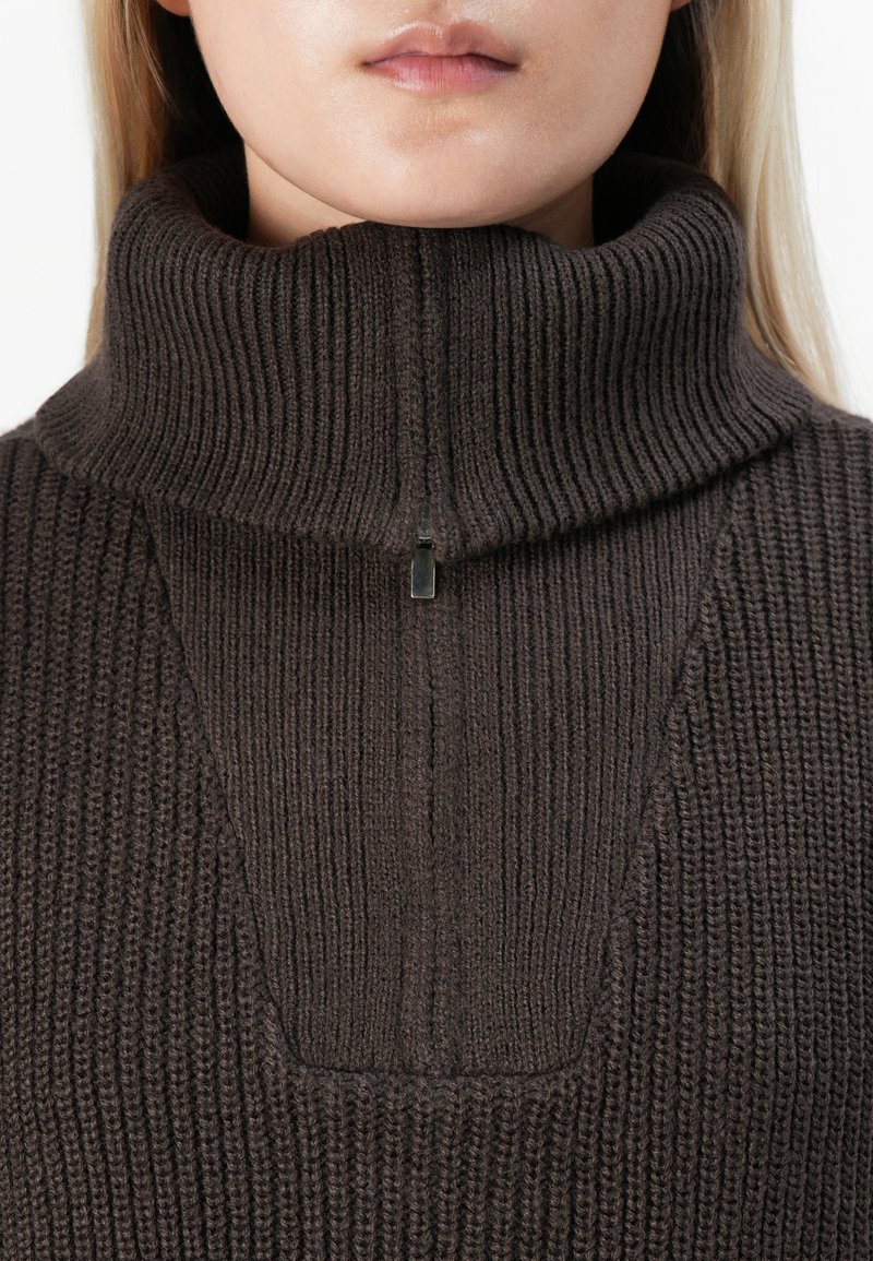 Gros plan sur une personne portant un pullover en tricot côtelé marron foncé avec un col montant zippé à moitié.