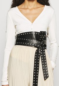 Una modella che indossa un top bianco a coste e una cintura larga nera con borchie argentate, abbinato a pantaloni beige plissettati.
