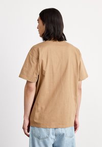 Brown cotton T-shirt with short sleeves, relaxed fit, crew neckline, and smooth texture; worn with light blue denim shorts.