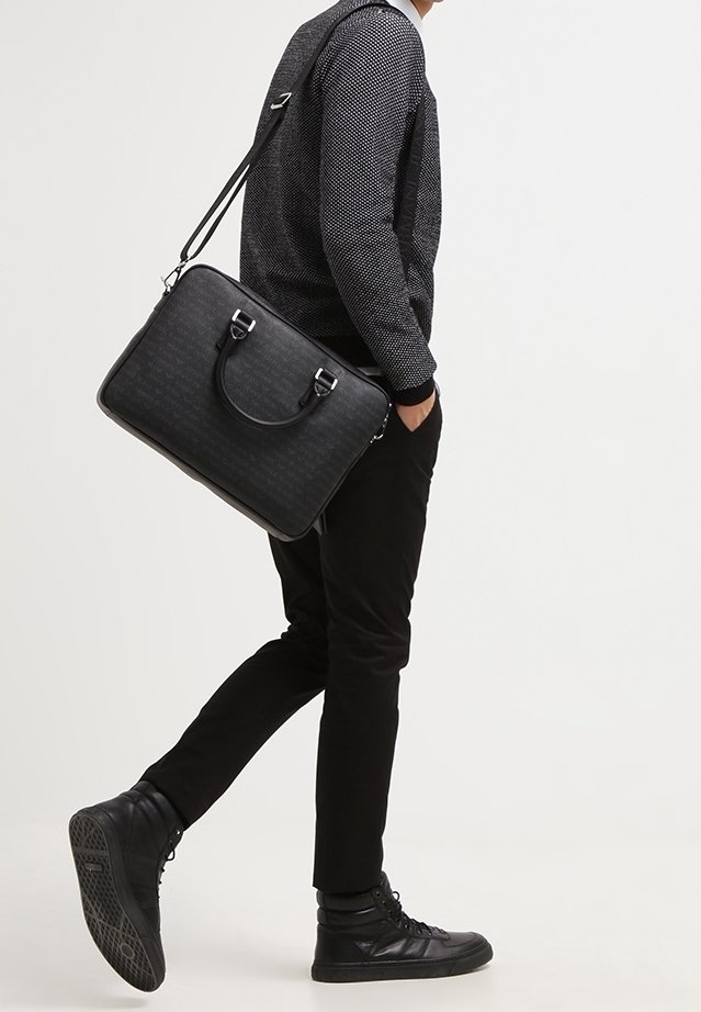 Personne marchant avec une main dans la poche, portant un pantalon noir, des baskets noires, un pull gris texturé, portant une mallette en cuir noire avec une bandoulière.