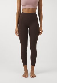 Leggings castanhos de cintura alta com uma textura suave, apresentando detalhes costurados. Acompanhados de um top de desporto rosa claro, sem padrões visíveis.