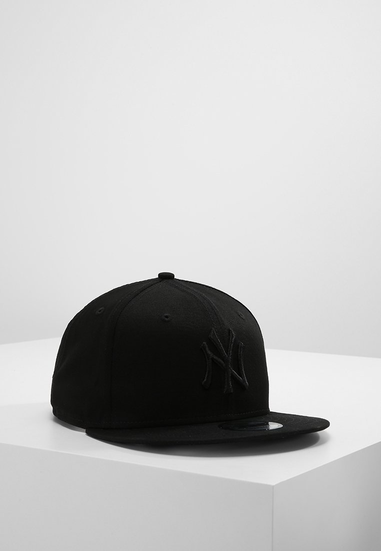 Casquette de baseball noire à visière plate avec logo en relief des New York Yankees à l'avant, exposée sur un socle blanc sur fond gris uni.