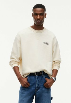 Junger Mann, der einen cremefarbenen Sweatshirt mit dem Text "Tommy", blaue Jeans, schwarzen Gürtel und silberne Uhr trägt, steht mit den Händen in den Taschen.