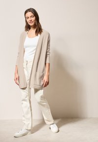 Ljusbeige stickad cardigan med långa ärmar och framfickor, buren över en vit linne och matchad med ljusa byxor och vita sneakers.