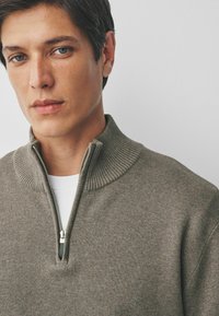 Close-up van een jonge man met blauwe ogen die een grijze half-zip sweater over een witte shirt draagt, tegen een effen lichte achtergrond.