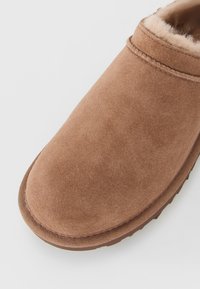 Lähikuva ruskeasta nubukista valmistetusta tossusta, jossa on pehmeä, pörröinen vuori ja kumipohja valkoisella taustalla.