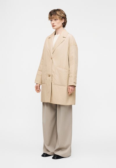 Γυναίκα όρθια φορώντας μπεζ φαρδιά oversized σακάκι, φαρδιά ανοιχτόχρωμα παντελόνια, μαύρα παπούτσια και χρυσούς κρίκους σκουλαρίκια σε λευκό φόντο.