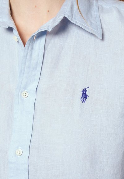 Hellblaues Leinenhemd mit Kragen, ausgestattet mit einem kleinen lila gestickten Logo und weißen Knöpfen. Weiche Textur, lässiges Design.