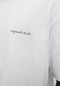 Camiseta de manga corta blanca hecha de algodón. Presenta texto en negro en forma de script "originals studio" en la parte izquierda del pecho. Textura suave.