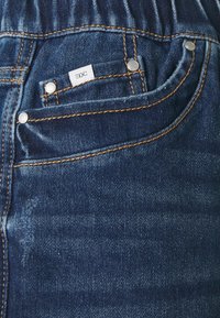 Sininen farkutasku, jossa on pieni valkoinen EDC-tagi, oranssia ommelta ja metallinitteitä taskun reunoilla.