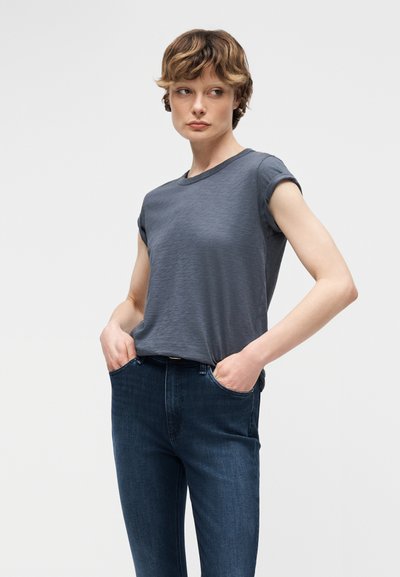 Άτομο με κοντά κυματιστά μαλλιά φορά γκρι κοντομάνικο μπλουζάκι με γυρισμένο μανίκι και σκούρο μπλε τζιν, στέκεται με τα χέρια στις τσέπες μπροστά σε απλό φόντο.