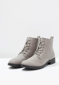 Stivaletti alla caviglia in suede grigio con suole nere piatte, lacci frontalmente, dettagli di cucitura e una cerniera laterale per una facile vestibilità.