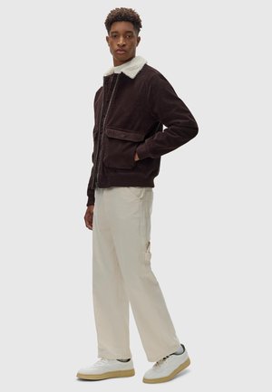 Donkergroene corduroyjas met een crème bontkraag, ritsluiting, dubbele vo zakken, gecombineerd met lichtbeige broek en witte sneakers.