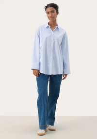 Camicia button-up a righe blu e bianche con maniche lunghe, abbinata a jeans blu a gamba dritta e sneakers bianche.