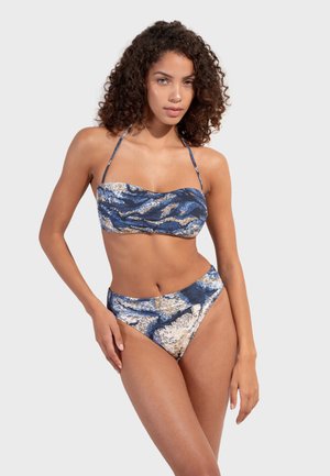 Kobieta w niebiesko-beżowym bikini bandeau z wzorem i paskami wiązanymi na szyi, stojąca na jednolitym szarym tle.