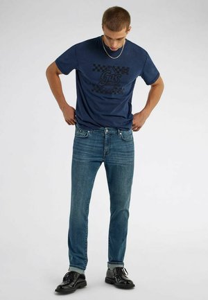 Junger Mann richtet ein blaues bedrucktes T-Shirt aus, kombiniert mit schlanken blauen Jeans und schwarzen Lederschuhen vor einem schlichten hellen Hintergrund.