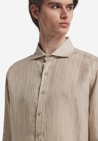 Camicetta beige a righe realizzata in tessuto leggero, con colletto a punta e chiusura frontale con bottoni di colore chiaro.